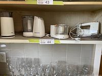 Shelf with the lot of four small electric kitchen appliances shown together and arranged in a row: Braun coffee grinder, Hamilton Beach mini chopper, Black & Decker mini chopper, Black & Decker hand blender with beaters.