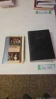 Two vintage books side by side on a white table: 1947 Winston dictionary (black hardcover) and 1962 opera themes and plots (colorful collage paperback with tape on spine)