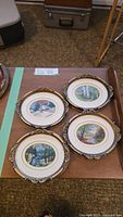 Set of four round needlepoint pictures in gold decorative frames showing various seasons in nature, arranged on table.