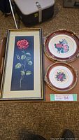 Three framed needlepoint floral art pieces arranged on a table; one rectangular with red rose, two round with floral patterns.