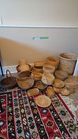 Photo showing collection of approximately twenty-one wicker baskets of various shapes and sizes arranged on a patterned rug against a wall.