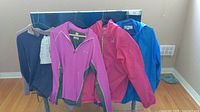 Four women's tops hanging with various colors and materials: pink Columbia half zip top, blue rain jacket, fuchsia nylon jacket, two-tone wool top.