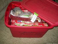 Red storage tub with lid partially open showing assorted gift supplies inside including a wrapped wreath or decoration.