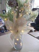 Photo of holiday floral center piece with white and green artificial foliage, glittery ornaments, and multi-colored battery operated string lights illuminated yellow.