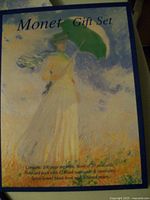Photo of the gift set box cover showing artwork of a woman in a white dress holding a green parasol with blue sky and orange grass background, listing contents as 100-page book, 20 postcards, and 10 blank notecards with envelopes.