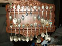 Front view of 24 silver souvenir spoons on wooden wall plaque, showing different crests and shapes of spoons including some decorative forks and scalloped bowls.