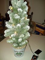 Photo showing tabletop Christmas tree with flocked branches, gold ornaments, warm white lights, and 'Farm Fresh Christmas Trees' decorative base container.