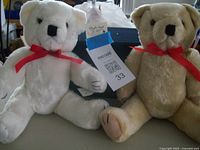 Two teddy bears, one white and one beige, each with red bows around their necks shown seated on a surface with maxsold tag attached to white bear.