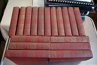 Seventeen red hardcover books arranged in two rows showing spines with gold text and embossed patterns.