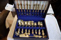 Open cutlery box showing arranged 24 kt gold plated knives, forks, spoons and extra serving pieces in blue velvet lining