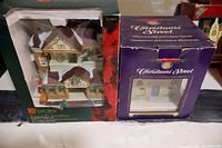 Two Christmas village houses in original boxes. One is a tan and brown house with Christmas decorations visible through plastic. The other is a blacksmith shop in purple box labeled Christmas Street.