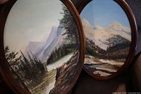 Two large oval oil paintings side by side showing forest and mountain landscapes in walnut frames.