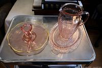 Displays the set of three pink Depression glass pieces on a metal surface: a jug, an oval tray, and a round tidbit tray with handle.