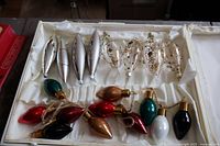 Box of assorted glass Christmas tree ornaments arranged within a lined box, including clear teardrop shapes, silver bullet shapes, and various colored bulb shapes.