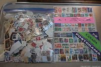 Large plastic bag filled with loose Canadian stamps alongside two unopened packs of Canadian stamps with colorful designs.