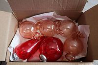 6 large pink and red glass Christmas ornaments displayed inside a cardboard box on tissue paper, showing 4 pink elongated bulbous ornaments and 2 red round ornaments.