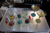 Shot showing six martini glasses with colored bases placed on a clear glass tray alongside two taller artist-signed martini glasses with painted designs.
