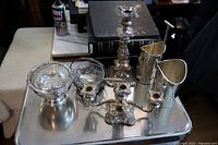 Full lot of silverplate items including candle holders, bowl, serving dish and spoons, and wine bottle holder arranged on metal tray.