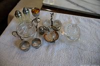 Lot of mixed silverplate and glass items displayed on white towel, including salt and pepper shakers, glass serving dish, napkin rings, small containers, and decorative plates.