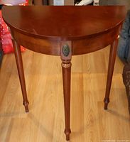 Front view of small demilune wooden table showing three legs, semi-circular top, decorative metal medallion, wood grain, and surface scratches.