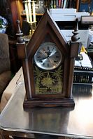 Front view of a wooden steeple clock with pointed top and finials, showing the clock face with Roman numerals and etched decorative glass panel below.