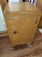 Closed view of a birds eye maple nightstand with visible wood grain and single door with black knob, showing wear and scuffs on top surface and sides.