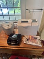 All items displayed on wooden table by window: two breadmakers and one waffle press, power cords visible, showing general condition and size.