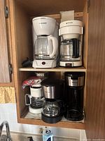 Four drip coffee makers arranged on wooden shelving, including Black & Decker and Betty Crocker brands, showing signs of wear
