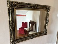 Full view of the large rectangular mirror hung on a wall in a room with wooden floors and chairs reflected in the glass.