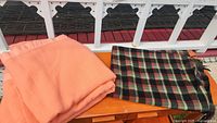 Photo showing the peach-colored Kenwood wool blanket alongside the multicolor tartan patterned blanket placed on a wooden surface outdoors.