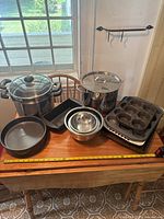 Full view of the kitchenware lot including pots, cake pan, mixing bowls, muffin tins, and baking trays on a wooden table with measuring tape for scale.