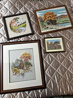Four framed textile artworks arranged on a quilted surface showing rustic landscapes and autumn scenes produced by needlepoint and crewel embroidery.
