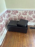 Black leather rectangular hassock ottoman with segmented cushioned removable top, showing wear and placed against wall with patterned wallpaper.
