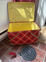 Open tin picnic basket showing yellow interior and signs of age, with a perforated skillet placed in front.