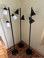 Three pedestal lamps shown together in corner with wood floor and wall background. Black metal poles and bases with white and black lamp heads.