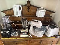 Full lot displayed on wooden furniture including food processor, blender, kettle, mixer, toaster, and rice cooker, showing interconnected cords and overall wear