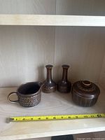 Four piece brown glazed ceramic set including a creamer, two vases, and a covered jar on a wooden shelf with measuring tape for scale