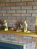 Photo showing four wood carvings: two hunter figures, one boy figure, and one beaver figure, arranged on a mantelpiece with measuring tape for scale.