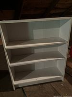 Front view of a white wooden bookcase with three shelves, placed in an attic setting with visible signs of wear and age.