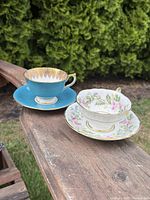 Two porcelain tea cups and saucers placed outdoors on a wooden surface with greenery background, showing colors and patterns of each set.