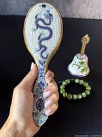 Hairbrush with blue dragon cloisonne design and floral patterns held in hand with bell and bracelet in background on black surface