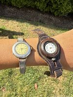 Two antique ladies wrist watches worn on wrist outdoors, one with a metal case and narrow strap, the other with large leather cuff wristband.