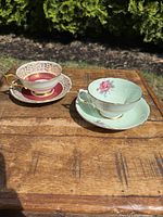 Two porcelain teacup and saucer sets placed on wooden surface outdoors with greenery in background.
