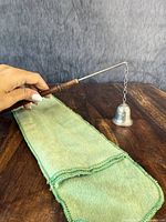 Photo showing full length of candle snuffer with wood handle and silver bell end, alongside green cloth pouch, with hand holding handle for scale