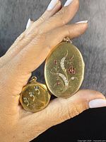 Both lockets front view held between fingers showing oval and octagonal shapes and gemstone designs