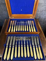 Full view of the open oak canteen box containing two rows of knives and forks with cream-colored carved handles and engraved metal blades and tines, resting on blue lining.