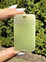 Front view of rectangular green carved jade plaque pendant held outdoors showing detailed top edge carvings and bail.