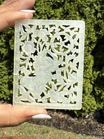 Front view of rectangular carved pierced jade plaque showing Foo Dog and floral motifs, held by hand outdoors.