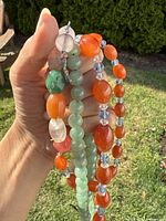 Hand holding four separate necklaces composed of various polished stones in orange, green, clear, and blue shades, outdoors on a lawn background.