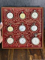 Overhead view of red velvet-lined wooden tray containing nine pocket watches